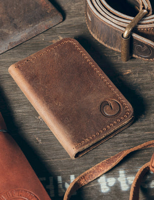 Brown leather wallet on a wooden surface with a belt and strap.